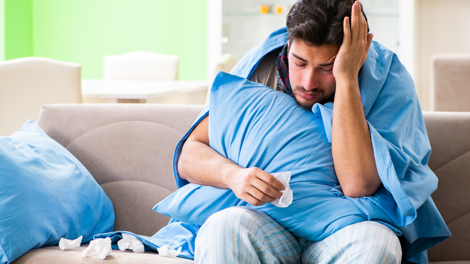 An ill man sitting on a couch with a blanket holding a pillow, with tissues, holding his head
